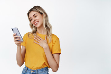 Portrait of touched young woman fond of something on smartphone screen, reading message on mobile phone with delighted smile, white background