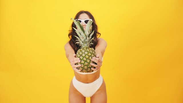 A positive brunette woman wearing a swimsuit giving an pineapple to the viewer standing isolated over a yellow background in the studio