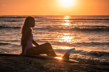 Fototapeta na wymiar Portrait of young blonde surfer girl with white long surf board single fin on the beach at sunset time