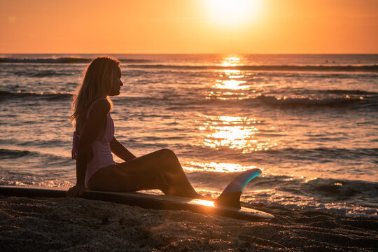 Portrait of young blonde surfer girl with white long surf board single fin on the beach at sunset time