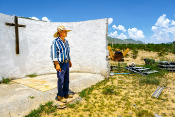 Full length of senior male rancher wearing hat looking away while standing against cross on white wall during sunny day