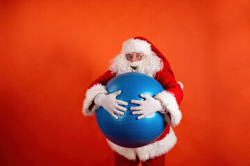 Funny fat Santa Claus with a rubber fitness ball on an orange background. Active sports and weight...