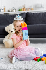 girl in toy crown showing tower of multicolored building blocks while sitting on floor