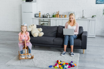 girl in costume of princess riding rocking horse near mother working on laptop on sofa - Powered by Adobe