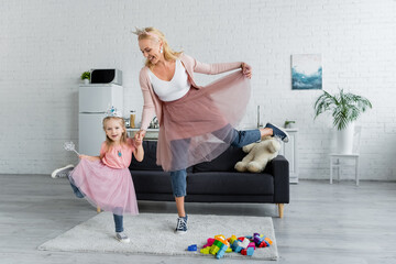 joyful mom and daughter in toy crowns and tulle skirts dancing at home