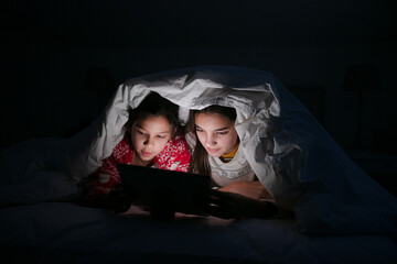 Sisters watching movie on digital tablet under blanket in dark bedroom