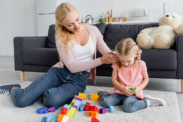 Naklejka premium smiling woman adjusting hair of daughter playing with building blocks on floor at home