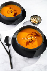 Two deep black bowls with an orange pumpkin cream soup with pumpkin seed oil, two black spoons on a pink marble surface