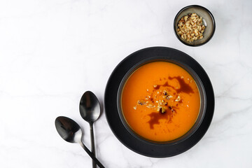 A deep black bowl with an orange pumpkin cream soup with pumpkin seed oil, two black spoons on a marble surface