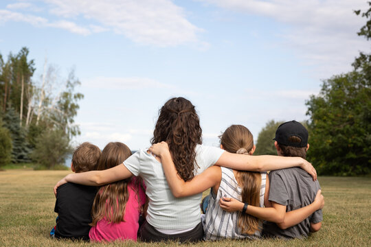 Group Of Youth Sitting On Grass Seen From Behind, Arms Around Each Other, Looking At View, With Copy Space In Sky