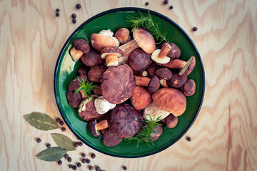 Freshly picked mushrooms laid out in a green bowl. Noble boletus and bay boletes surrounded by herbs on a plywood table.