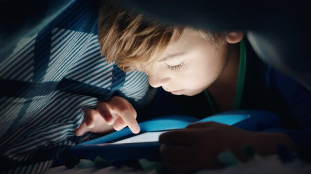 happy little boy using digital tablet computer under blanket enjoying learning on touchscreen technology playing games having fun at bedtime