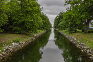 A narrow canal with paved banks. The canal is surrounded by large deciduous trees that are reflected in the water surface