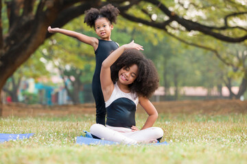 Naklejka na ściany i meble Smiling African American little girl practicing yoga and stretching her hands on exercise mat outdoor in the park. Two little afro girl with curly hairstyle training yoga