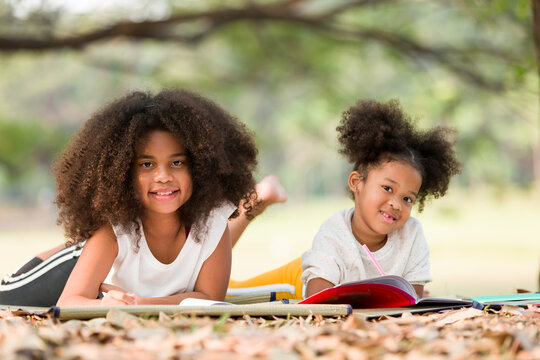 Happy two African American little kids girl drawing on notebook with pencil while lying on mat outdoor. Two little girl using pencil writing and doing homework on notebook together in the park