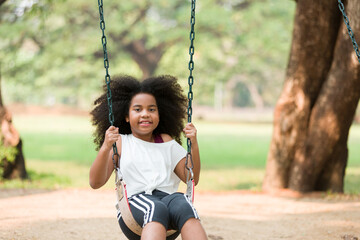 Happy cheerful African American child girl playing on swing at playground in park. Smiling girl having fun on a swing