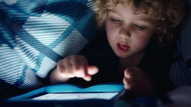 happy little boy using digital tablet computer under blanket enjoying learning on touchscreen technology playing games having fun at bedtime