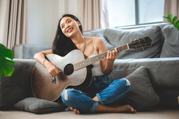 Asian woman playing music by guitar at home, young female guitarist musician lifestyle with acoustic art instrument sitting to play and sing a song making sound in hobby in the house room - obrazy, fototapety, plakaty