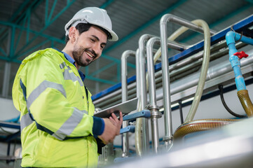 Young male machine inspector wearing vest and hardhat with headphones checking machine and sterilizers in water plant while making notes in digital tablet