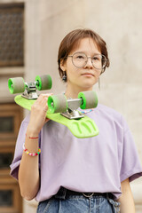 brunette teen girl with glasses with a green skateboard on the streets of the city on a clear sunny day