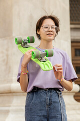 brunette teen girl with glasses with a green skateboard on the streets of the city on a clear sunny day