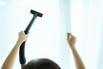 Asian young woman using vacuum machine to cleanup the dust on curtain in living room close up. Housework and chores in daily life concept. Housekeeper cleaning a white curtain.