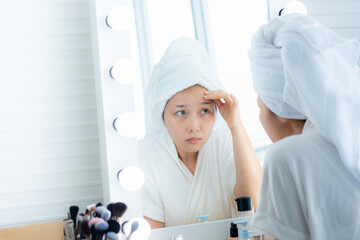Asian woman in a bathrobe and covering her hair with a towel have a serious face Look in the mirror and see acne. Wrinkles and dark spots on the face