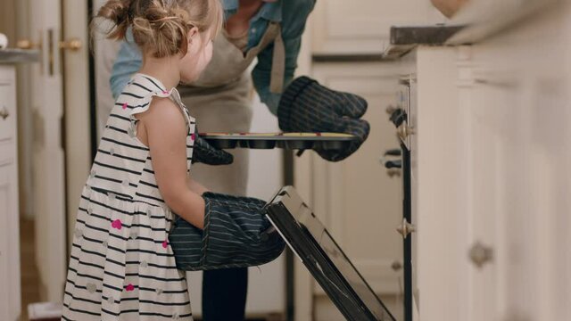 happy little girl helping mother bake in kitchen putting homemade cupcakes in oven wearing oven mitts enjoying fresh delicious treats