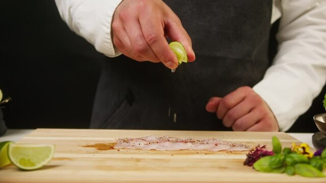 Professional Chef squeezing lime juice on raw white fish fillet on cutting board close-up. Man cooking seafood dishes using citrus fruits. Process of preparing ingredients for Peruvian ceviche. 