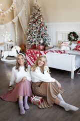 Two happy young women friends in cozy sweaters adorn the Christmas tree laugh give gift boxes for the New Year holiday in the decorated interior of the bedroom of the house, selective focus