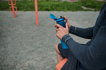 Close-up of an unrecognizable athlete holding a fresh water bottle rehydrating his body after exercise, bodybuilding and outdoor workout. Cropped image
