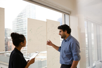 Businessman and businesswoman discussing diagram sketches hanging on conference room window