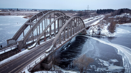 winter, iron bridge for cars and trains, over a frozen river, transparent ice, top view, drone, blue shades, the sky is blue, buildings can be seen here and there