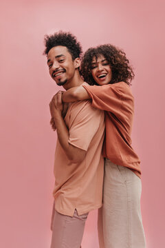 profile photo of young mulatto woman hugging from behind tall guy indoors in pink. happy brunettes with curly hair are dressed in light summer clothes.