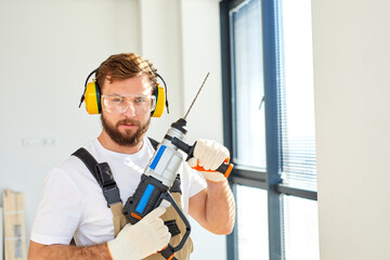 Fototapeta premium Young caucasian man holding power drill standing in front of white wall indoors,posing at camera, wearing overalls, headset and safety glasses. copy space, concept for home DIY
