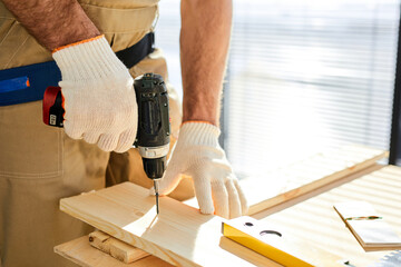 Woodworker drills hole with an electric drill in wood board. Close-up photo of hands with drill, side view. repairing, carpentry, housework, construction, woodworking concept