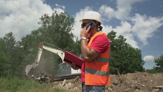 Male engineer wearing helmet and vest talking by phone at construction site against working excavator