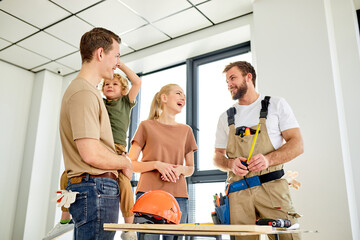 Cheerful Family With Kid Boy Having Talk With Friendly Handyman, Discussing House Layout, Home Renovation. Man And Woman With Child Boy And Builder In Overall Smile While Talking