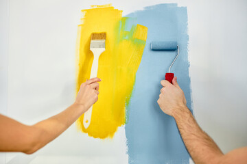 Young couple painting walls in new house, using paint roller, brush, close-up photo of hands, cropped man and woman renovate home interior together. copy space