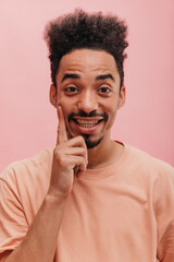 image of young happy amazed African American guy with amazing facial expression. dressed in peach-colored T-shirt, looks into camera touching his cheeks, stands on pink background.