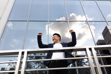 Asian businessman boss stands near his office center on the balcony in a dark color business suit holds his hands up celebrates victory