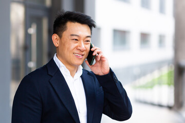 portrait Asian businessman talking on the phone cheerful smiling reports good news near the office center outside looking at the camera