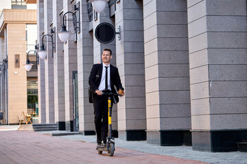Joyous caucasian business man in formal suit riding an electric scooter by city building after business meeting, holding cup of coffee in hands, take a break. Ecological transportation concept. © Roman