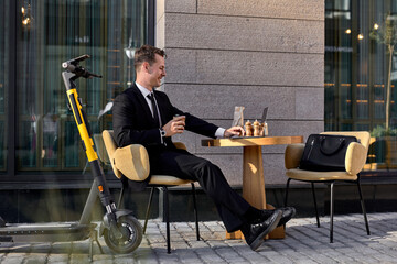 Happy business man in formal suit sit working on laptop in outdoor cafe, an electric scooter standing by him, handsome guy in formal wear sit drinking coffee, enjoying freelance. side view portrait