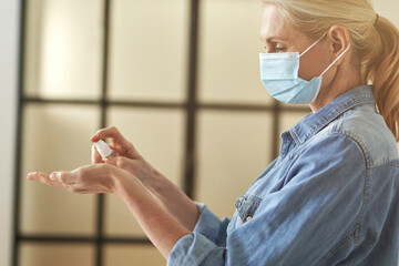 Side view of mature blonde woman in protective face mask cleaning her hands using antibacterial hand sanitizer gel pump dispenser, standing indoors