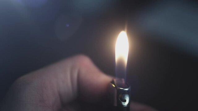 A gas lighter in a man's hand lights up in slow motion. A flame of fire illuminates a dark room, burns with sparks on a black abstract background.