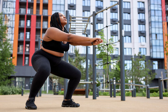 Physical exercise squat for fitness performed by obese african young woman, body positivity and healthy lifestyle concept. Side view on focused lady in black sportive wear looking at side. copy space