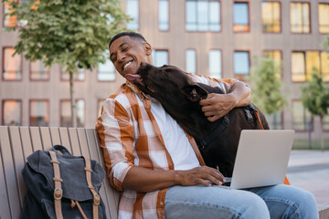 Happy African American man freelancer using laptop computer, working online hugging with adorable pet. Labrador dog licking his owner in park, selective focus. Best friends concept  - obrazy, fototapety, plakaty