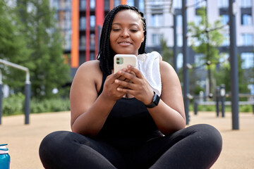 african woman sit in sports ground having rest chatting with friend on smartphone, typing message and smiling, enjoying sport, in black tracksuit, relax after workout. copy space. people lifestyle