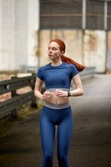 Fototapeta na wymiar Young slim redhead woman warming up on bridge before workout, running. Beautiful young fit fitness sport model jogging alone, side view. healthy lady in blue sportive clothes. motivation concept